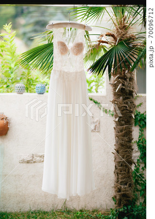 The bride's wedding dress with a lace transparent corset and a beige bodice on a hanger near a white stone fence and a decorative palm tree. The bride's wedding dress with a lace transparent corset and a beige bodice on a hanger near a white stone fence and a decorative palm tree. 70096712