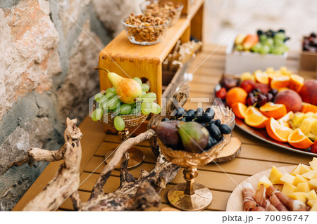 Gilded bowls with blue and white grapes, figs and pears on a wooden table with ham, cheese and fruits on a tray. 70096747