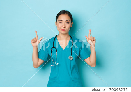 Covid-19, social distancing and coronavirus pandemic concept. Skeptical and doubtful young asian female doctor, professional physician or nurse in scrubs pointing fingers up, light blue background Covid-19, social distancing and coronavirus pandemic concept. Skeptical and doubtful young asian female doctor, professional physician or nurse in scrubs pointing fingers up, light blue background 70098235