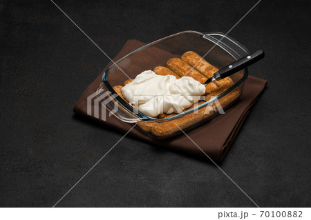 tiramisu dessert cooking - Traditional Italian Savoiardi ladyfingers Biscuits and cream in glass baking dish on dark concrete background 70100882