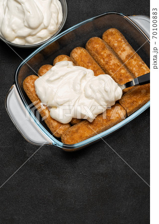 tiramisu dessert cooking - Traditional Italian Savoiardi ladyfingers Biscuits and cream in glass baking dish on dark concrete background 70100883