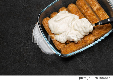 tiramisu dessert cooking - Traditional Italian Savoiardi ladyfingers Biscuits and cream in glass baking dish on dark concrete background 70100887
