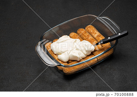tiramisu dessert cooking - Traditional Italian Savoiardi ladyfingers Biscuits and cream in glass baking dish on dark concrete background 70100888