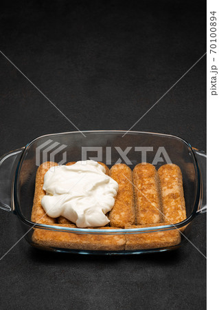 tiramisu dessert cooking - Traditional Italian Savoiardi ladyfingers Biscuits and cream in glass baking dish on dark concrete background 70100894