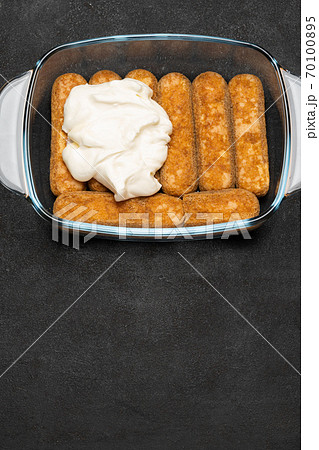 tiramisu dessert cooking - Traditional Italian Savoiardi ladyfingers Biscuits and cream in glass baking dish on dark concrete background 70100895