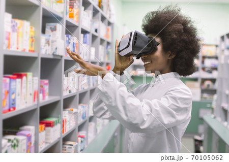 Side view of young professional African woman pharmacist, wearing virtual reality headset, working in modern drugstore and looking at shelves with medicines and gesturing 70105062