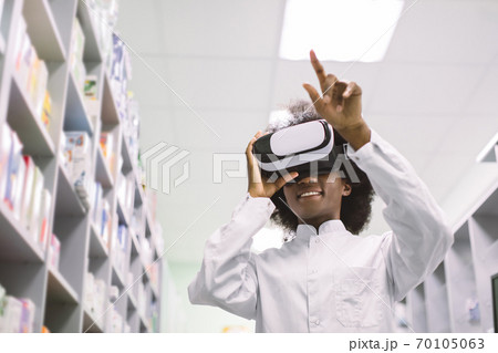 Bottom angle view of young pretty African American female clinical pharmacist or doctor, working in modern hospital drugstore in virtual reality goggles Bottom angle view of young pretty African American female clinical pharmacist or doctor, working in modern hospital drugstore in virtual reality goggles 70105063