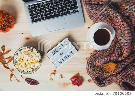 Top view Movie night concept. Flat lay composition with Movie night message on the board, laptop, popcorn bowl, decorative pumpkin, fallen leaves, a cup of tea, and warm plaid on wooden background. Top view Movie night concept. Flat lay composition with Movie night message on the board, laptop, popcorn bowl, decorative pumpkin, fallen leaves, a cup of tea, and warm plaid on wooden background. 70106602