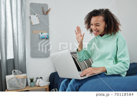 Young cheerful mixed-race woman waving hand to her friend on laptop display Young cheerful mixed-race woman waving hand to her friend on laptop display 70118584