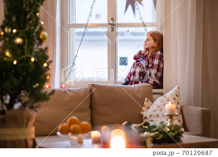 Portrait of small sad girl in pajamas indoors at home at Christmas, sitting on window. 70120687