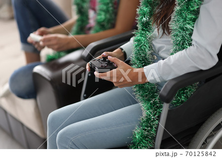 Disabled woman in wheelchair and tinsel around neck holds joystick from computer console with friend at home 70125842