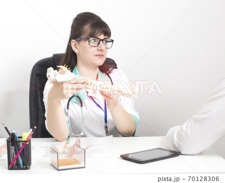 A female orthopedic doctor tells the cause of back pain in a patient using an example of a spine layout. Scoliosis and facet joint syndrome, copy space A female orthopedic doctor tells the cause of back pain in a patient using an example of a spine layout. Scoliosis and facet joint syndrome, copy space 70128306