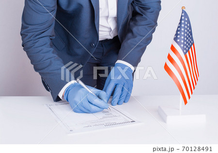Man in protective medical gloves votes on presidential election of USA during coronavirus pandemic COVID 19. Person fills out paper ballot. Flag of the United States is on the table. General voting 70128491