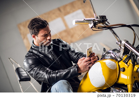 A beautiful muscular biker leads a video blog and talks on a video call on a smartphone while sitting on a motorcycle in the garage 70130319