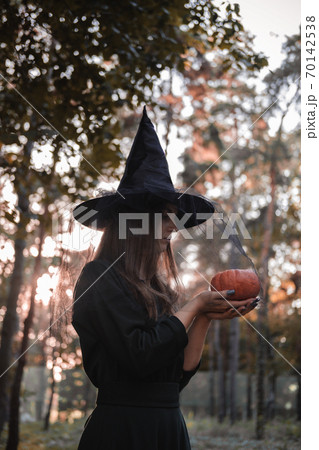 Young cute beautiful woman in dark dress and witch's hat holds an orange pumpkin in her hands. Halloween party costume. Forest, park with autumn trees. Young cute beautiful woman in dark dress and witch's hat holds an orange pumpkin in her hands. Halloween party costume. Forest, park with autumn trees. 70142538