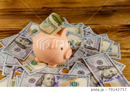 Pink piggy bank on a heap of american one hundred dollars bills on a wooden desk Pink piggy bank on a heap of american one hundred dollars bills on a wooden desk 70142757