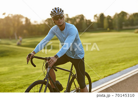 Strong athletic man in sportswear and protective helmet standing with his bicycle on the road in park and looking away, resting after cycling outdoors Strong athletic man in sportswear and protective helmet standing with his bicycle on the road in park and looking away, resting after cycling outdoors 70145310