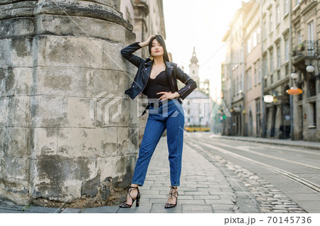 Full length portrait of young brunette Asian woman in jeans and stylish black leather jacket, sexy posing near old stone wall of the building, outdoor summer shot 70145736