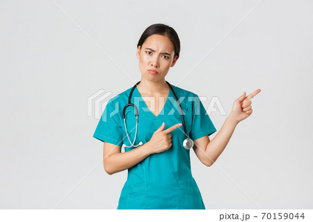 Covid-19, healthcare workers, pandemic concept. Disappointed and angry asian female therapist, nurse in scrubs looking upset at camera, sulking and pointing upper right corner, wait explanation Covid-19, healthcare workers, pandemic concept. Disappointed and angry asian female therapist, nurse in scrubs looking upset at camera, sulking and pointing upper right corner, wait explanation 70159044