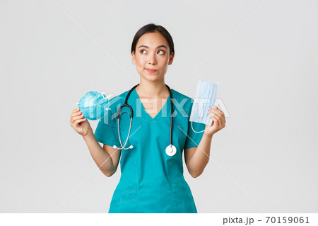 Covid-19, coronavirus disease, healthcare workers concept. Thoughtful smiling asian female doctor, physician in scrubs looking dreamy upper left corner, holding respirator and medical mask Covid-19, coronavirus disease, healthcare workers concept. Thoughtful smiling asian female doctor, physician in scrubs looking dreamy upper left corner, holding respirator and medical mask 70159061