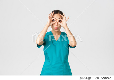 Covid-19, healthcare workers and preventing virus concept. Impressed funny and cute female asian nurse in scrubs looking through fingers over eyes as if stare in binoculats amazed 70159092
