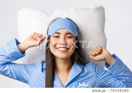 Close-up of happy and pleased smiling asian girl in blue pajamas and sleeping mask lying in bed on pillow, stretching satisfied and grinning after having good night sleep, waking up energized Close-up of happy and pleased smiling asian girl in blue pajamas and sleeping mask lying in bed on pillow, stretching satisfied and grinning after having good night sleep, waking up energized 70159160