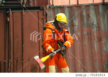 A portrait of Asian male fireman in orange protective clothing, mask and helmet with an ax standing front of building. A portrait of Asian male fireman in orange protective clothing, mask and helmet with an ax standing front of building. 70163914