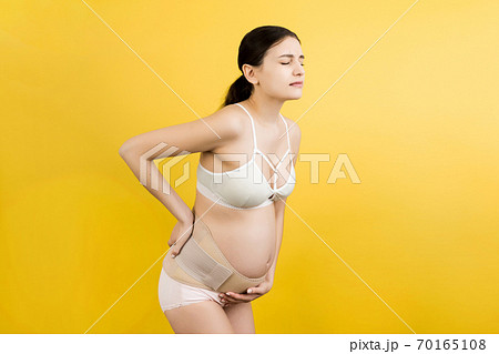 Portrait of pregnancy belt dressed on suffering pregnant woman in underwear for reducing pain in the back at yellow background with copy space. Orthopedic abdominal support belt concept Portrait of pregnancy belt dressed on suffering pregnant woman in underwear for reducing pain in the back at yellow background with copy space. Orthopedic abdominal support belt concept 70165108