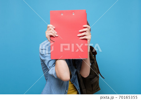 Caucasian student hiding her face behind a book, isolated on blue background. 70165355