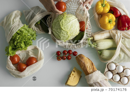 Woman came back from a market and unpacks a reusable grocery bag full of vegetables on a kitchen at home. Zero waste and plastic free concept. Girl is holding mesh cotton shopper with vegetables. 70174176