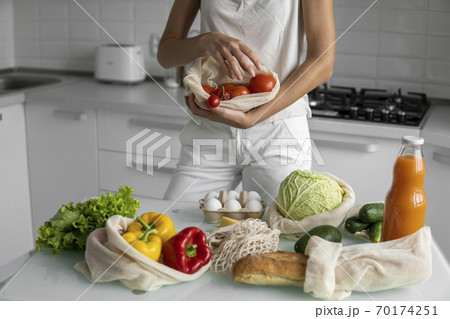 Woman's hand, holding a reusable grocery bag with vegetables on a kitchen at home and takes tomato out. Zero waste and plastic free concept. Mesh cotton shopper with vegetables. Ecology. 70174251