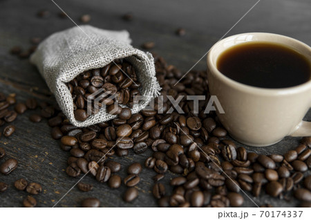 Coffee scattered fron a linen bag and a cup of black coffee Espresso or Americano standing on a wooden table. Fresh arabica coffee beans on linen textile. Aromatic freshly brewed coffee. Coffee scattered fron a linen bag and a cup of black coffee Espresso or Americano standing on a wooden table. Fresh arabica coffee beans on linen textile. Aromatic freshly brewed coffee. 70174337