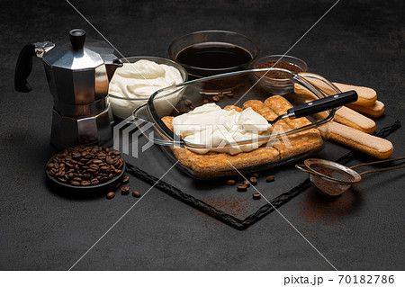 tiramisu dessert cooking - Traditional Italian Savoiardi ladyfingers Biscuits and cream in glass baking dish, coffe maker on dark concrete background 70182786