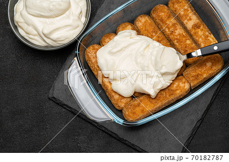 tiramisu dessert cooking - Traditional Italian Savoiardi ladyfingers Biscuits and cream in glass baking dish on dark concrete background 70182787