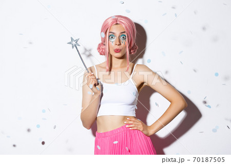 Attractive girl with magic wand celebrating halloween in fairy costume and pink wig, looking coquettish at camera, standing over white background while confetti falling down 70187505