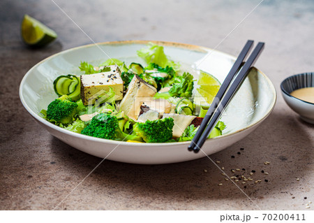 Green vegan salad with broccoli, smoked tofu and tahini dressing in white bowl, dark background. Green vegan salad with broccoli, smoked tofu and tahini dressing in white bowl, dark background. 70200411