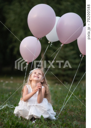 Little girl in white dress with balloons sitting on grass 70200588