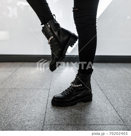 Young modern woman in black stylish jeans in fashionable leather lace-up boots stands in a store. Trendy collection of women's autumn shoes with rhinestones. Close up of female legs in footwear. 70202403