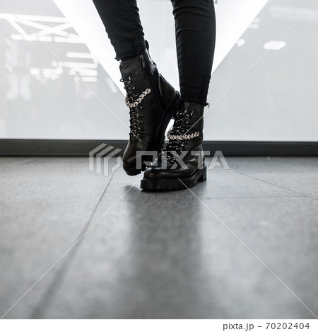 Fashionable women's seasonal shoes. New collection. Modern young woman in stylish black leather boots in vintage jeans stands in a bright room. Closeup of female legs in trendy footwear. 70202404