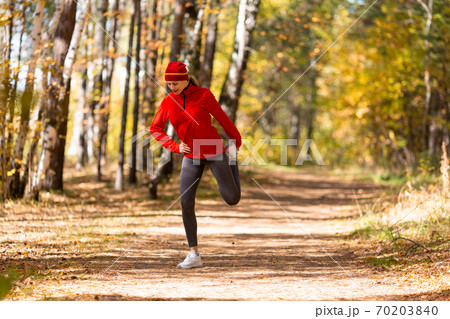 Legs warm up before jogging. Caucasian woman in red sportswear does hips stretching in the forest in autumn outdoor, selective focus 70203840