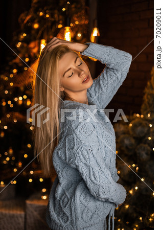 Young European model of a beautiful woman in a trendy blue knitted sweater posing in a dark room near a bright festive Christmas tree with vintage garlands. Sexy stylish sensual girl blonde indoors. Young European model of a beautiful woman in a trendy blue knitted sweater posing in a dark room near a bright festive Christmas tree with vintage garlands. Sexy stylish sensual girl blonde indoors. 70209011