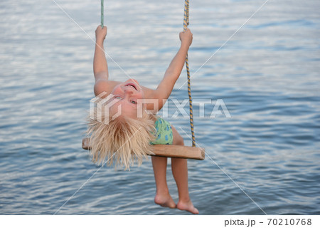 Fair-haired boy rides a rope swing on sea background. Happy childhood. 70210768