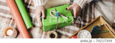 Young woman sits on plaid in cozy knitted woolen white sweater, socks and wraps Christmas gift in polka dot wrapping paper. Wooden tray with mug of cocoa with marshmallows, toy tree, candle, straws. Young woman sits on plaid in cozy knitted woolen white sweater, socks and wraps Christmas gift in polka dot wrapping paper. Wooden tray with mug of cocoa with marshmallows, toy tree, candle, straws. 70217304