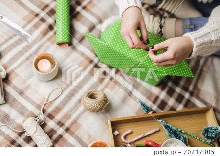 Young woman sits on plaid in cozy knitted woolen white sweater, socks and wraps Christmas gift in polka dot wrapping paper. Wooden tray with mug of cocoa with marshmallows, toy tree, candle, straws. 70217305