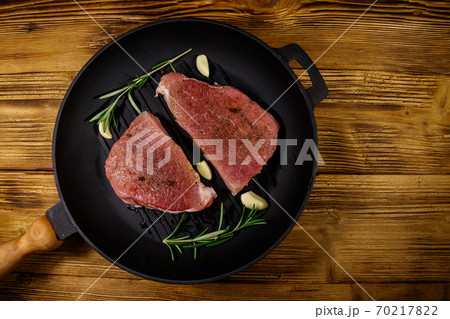 Raw pork steaks ready for preparation with rosemary, garlic and spices in cast iron grill frying pan on wooden table. Top view 70217822