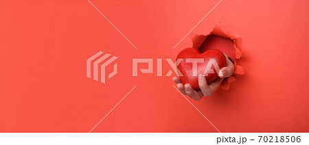 Hand holding red heart on torn red paper wall. Copy space aside for your advertising and offer or sale content. Valentines day, mother day, social advertising concept Hand holding red heart on torn red paper wall. Copy space aside for your advertising and offer or sale content. Valentines day, mother day, social advertising concept 70218506