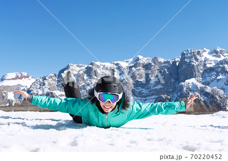 A teenage girl in a colorful ski suit, helmet and sunglasses lies in the snow and smiles happily against the backdrop of mountain peaks. Sports concept, people, travel. 70220452