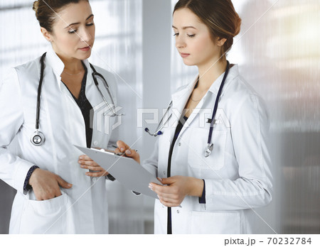 Two female physicians are discussing their patient's medical tests, while standing in a sunny clinic office. Doctors use a clipboard at work. Teamwork in medicine Two female physicians are discussing their patient's medical tests, while standing in a sunny clinic office. Doctors use a clipboard at work. Teamwork in medicine 70232784