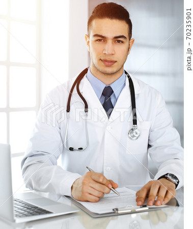 Arab doctor man is using laptop computer while filling up medication history records form sunny clinic. Medicine concept 70235901