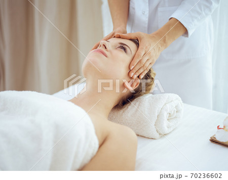 Young and blonde woman enjoying facial massage in spa salon. Beauty concept Young and blonde woman enjoying facial massage in spa salon. Beauty concept 70236602
